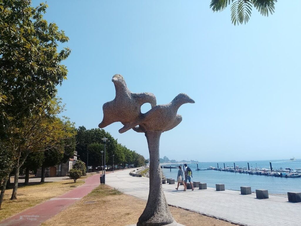 Escultura As Gaivotas junto al paseo marítimo de Cangas do Morrazo, con el mar turquesa y las playas de la ría de Vigo al fondo.