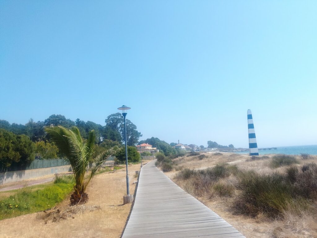 Pasarela de madera entre vegetación costera con un faro al fondo en Cangas do Morrazo.