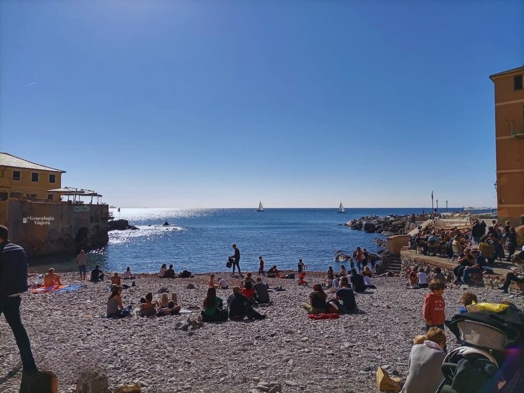 Playa de guijarros en Boccadasse, Génova, con turistas relajándose junto al mar Mediterráneo y veleros en el horizonte durante un día soleado de octubre en Liguria, Italia.