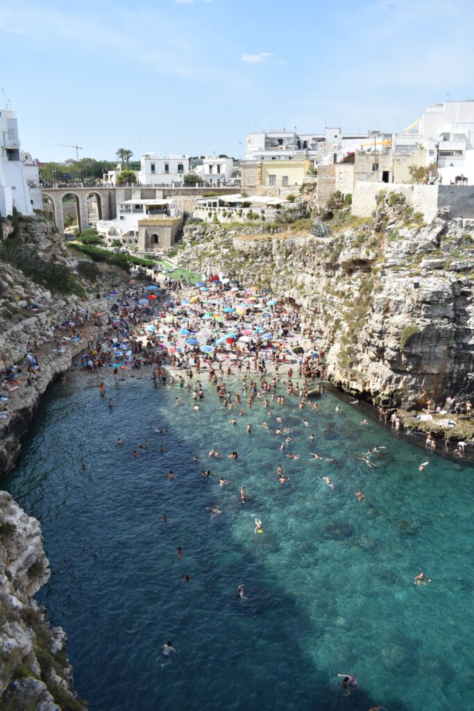 Vista desde el mar de Cala Porto o Lama Monachile en Polignano a Mare con el puente de piedra, los acantilados y visitantes disfrutando del mar Adriático