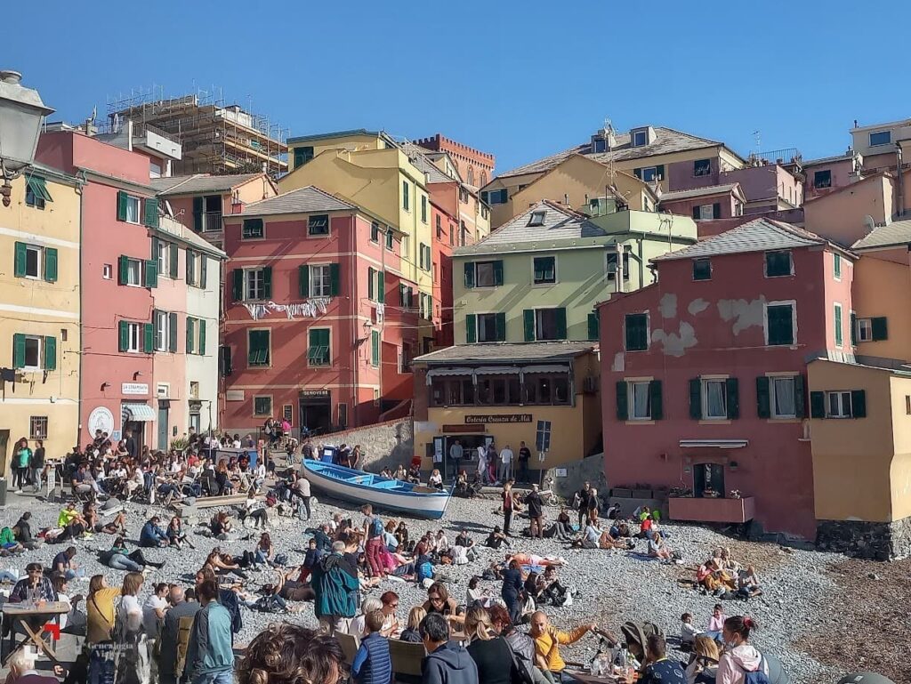 Casas de colores que rodean la playa de Boccadasse en Génova, con numerosos visitantes disfrutando de la costa de piedra en un día de verano.