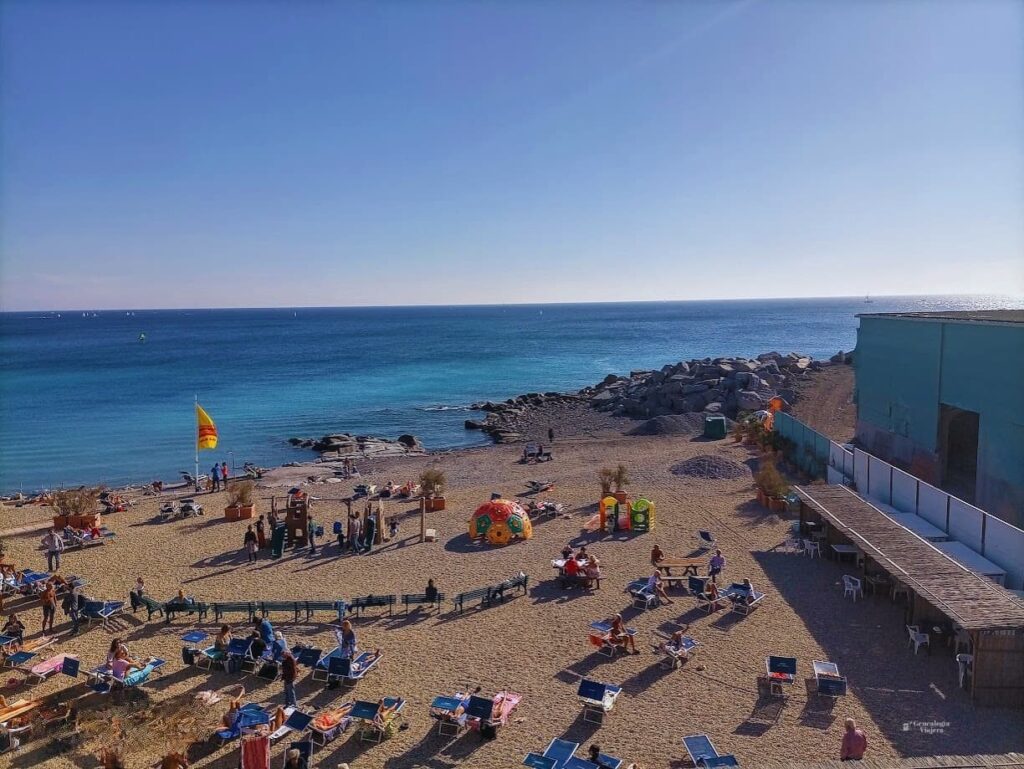 Playa de grava junto a Corso Italia en Génova, con personas descansando en reposeras y sombrillas frente al mar azul.