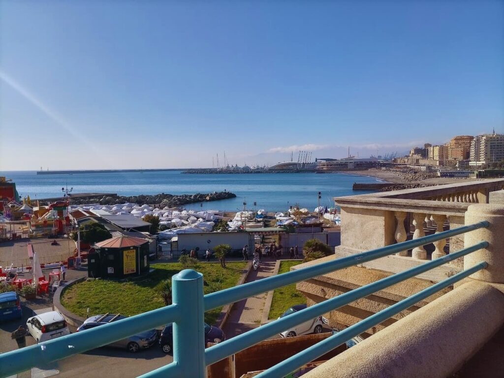 Panorámica de Corso Italia en Génova con la baranda del paseo, las playas con sombrillas, bares costeros con el mar y la ciudad de fondo.