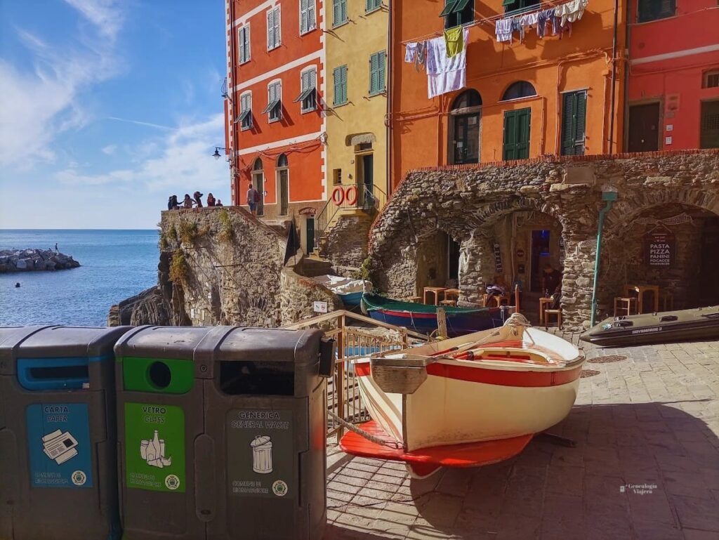 ista del pintoresco puerto de Riomaggiore en Cinque Terre, con botes pesqueros de madera en primer plano junto a botes de reciclaje, y edificios de colores cálidos sobre un acantilado rocoso con el mar Mediterráneo de fondo.