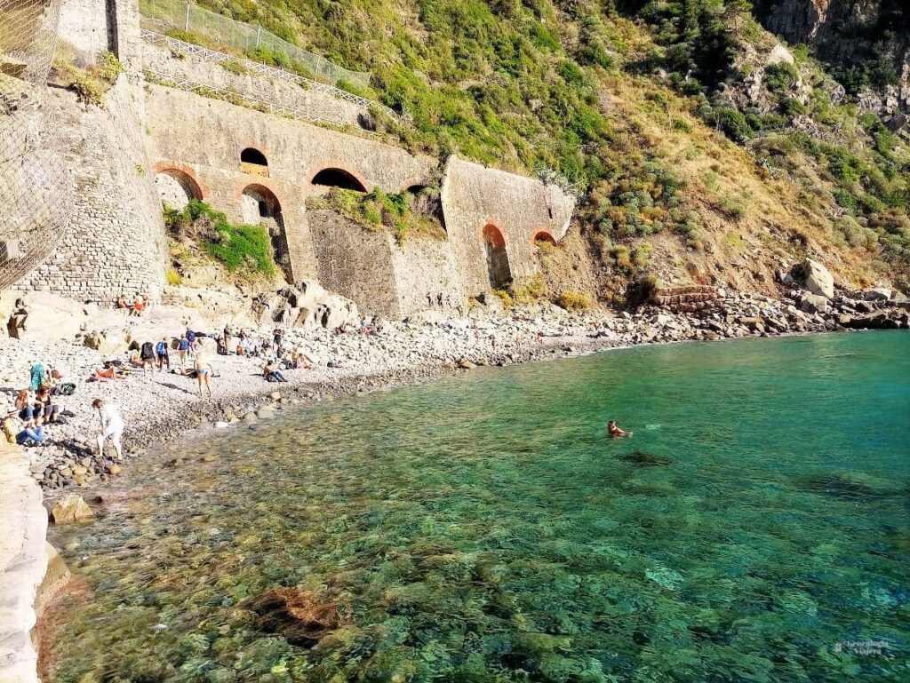 Playa de guijarros en Riomaggiore, Italia, mostrando el mar cristalino de tonos turquesa, personas descansando en la orilla y los antiguos arcos de piedra construidos sobre el acantilado