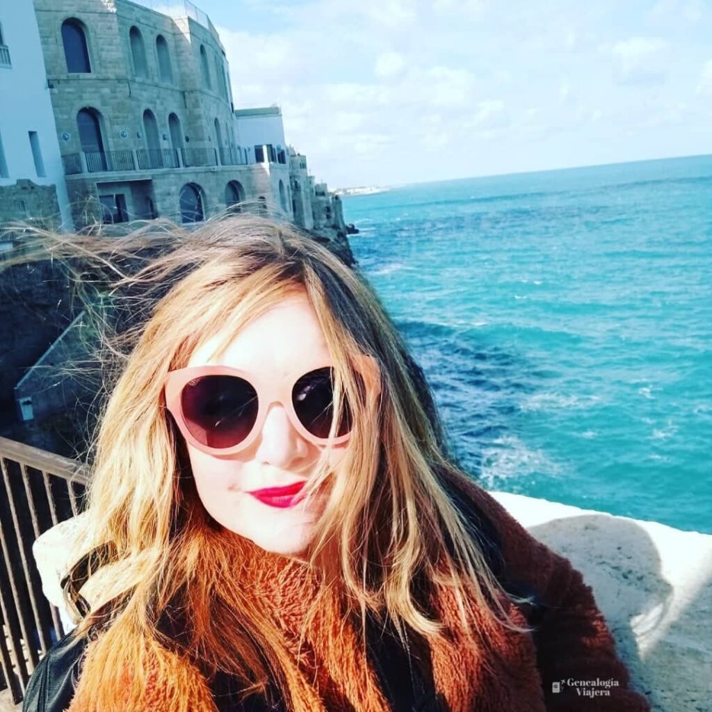 Viajera con abrigo en Polignano a Mare durante una visita en octubre, con el mar Adriático y la ciudad de fondo