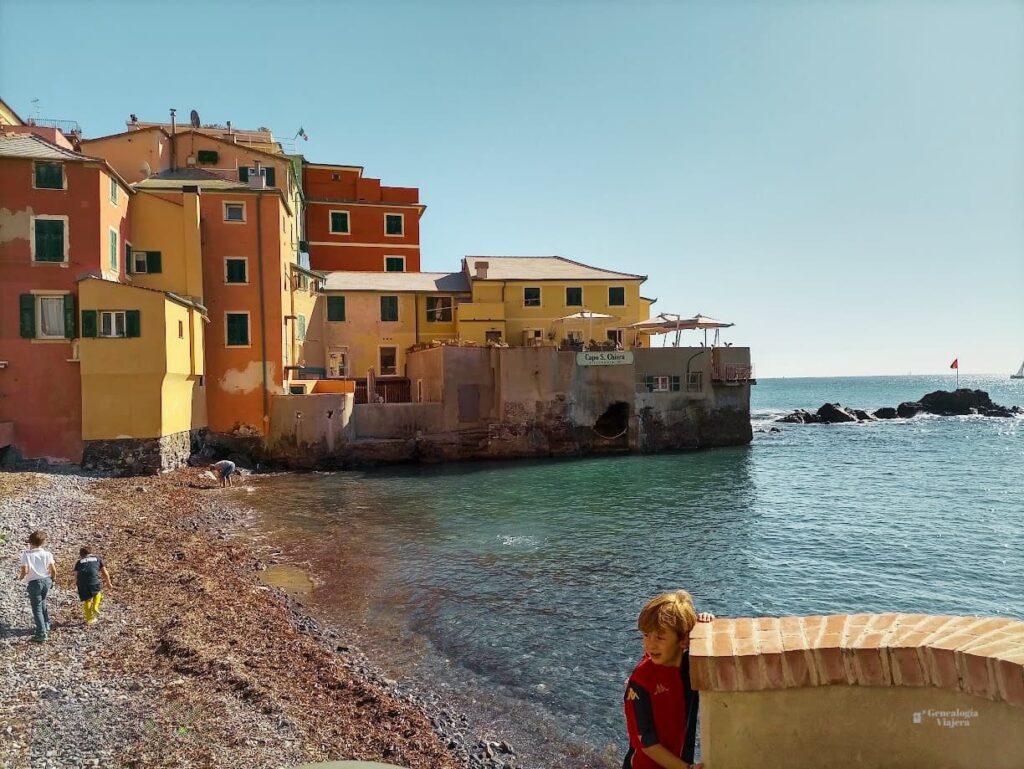 ista de la playa de Boccadasse en Génova, con las casas coloridas frente al mar, una terraza con mesas sobre el paseo y personas disfrutando de la playa.