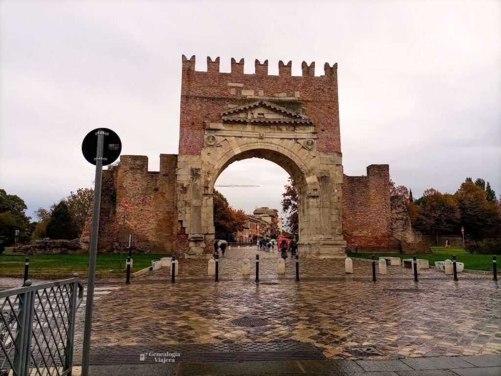 El antiguo Arco de Augusto en Rímini, una estructura romana de piedra y ladrillo, bajo un cielo nublado y con el suelo de adoquines mojado por la lluvia.