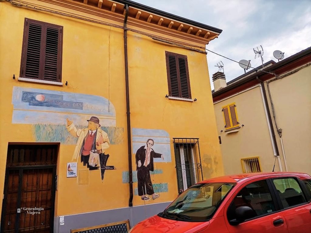 Frente de una casa amarilla en Borgo San Giuliano, Rímini, decorada con un mural del artista Agim Sulaj que rinde homenaje a la película 'La voce della luna' de Federico Fellini. En la pintura se ve a Roberto Benigni señalando al cielo. Un auto rojo está estacionado en primer plano.