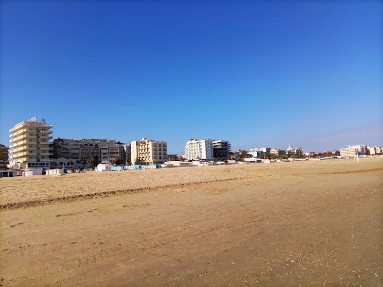 Panorámica desde la arena de la playa de Rímini mirando hacia la ciudad, mostrando grandes hoteles frente al mar sin turistas ni sombrillas.