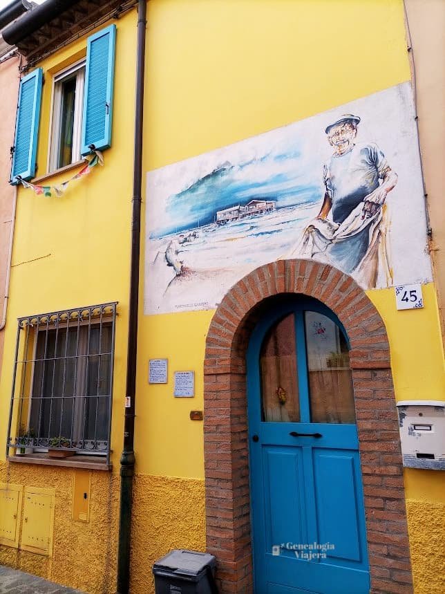 Fachada amarilla de una casa antigua en el Borgo San Giuliano de Rímini, Italia. Destaca una puerta de madera azul brillante bajo un arco de ladrillos y ventanas con persianas celestes. En la parte superior de la entrada, un mural pintado al estilo acuarela muestra a un viejo pescador recogiendo sus redes frente a un mar picado y una estructura de palafitos costeros.