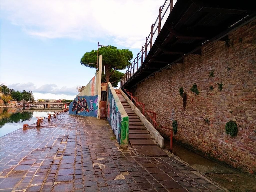 Paseo peatonal junto al río en Rímini, Italia, durante el mes de octubre. La imagen muestra un camino de adoquines húmedos que bordea el agua, un muro alto de ladrillo antiguo a la derecha con escaleras que suben hacia una plataforma, y paredes decoradas con grafitis coloridos bajo un pino y un cielo parcialmente cubierto.