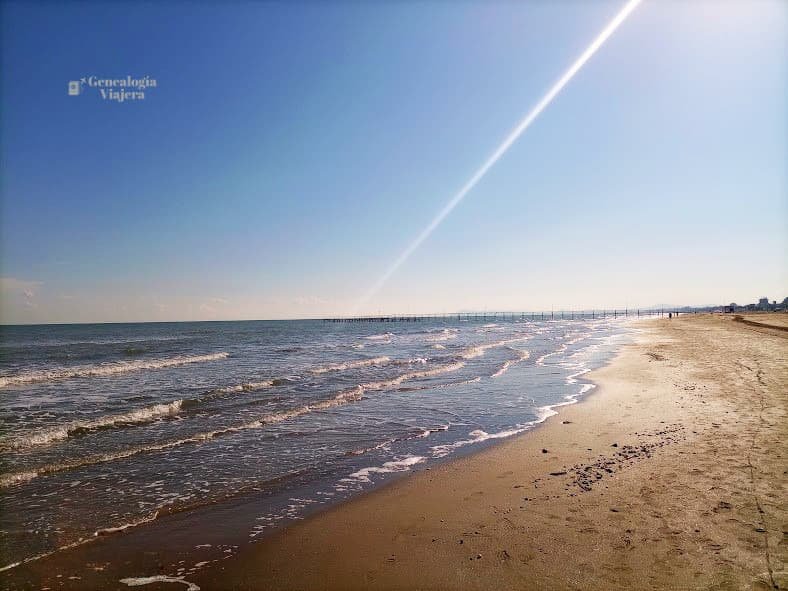 Orilla de la playa de Rímini con arena dorada húmeda y pequeñas olas con espuma, mostrando un muelle largo en el horizonte y un intenso reflejo de luz solar sobre el mar calmo.
