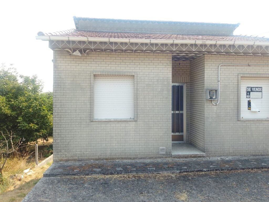 Fachada de vivienda unifamiliar con persiana bajada y cartel de se vende en la ventana en Sandiás junto a la ruta.