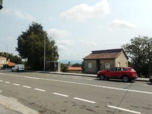 arada de autobús tipo marquesina junto a la carretera con coche rojo estacionado y viviendas al fondo en Sandiás.