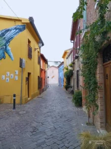 Una pintoresca calle peatonal empedrada en el Borgo San Giuliano de Rímini, Italia, durante el mes de octubre. La escena está enmarcada por coloridas fachadas residenciales de tonos amarillos, naranjas y azules, destacando a la izquierda un mural de una ballena pintado en una pared y plantas trepadoras en un muro de ladrillo a la derecha.