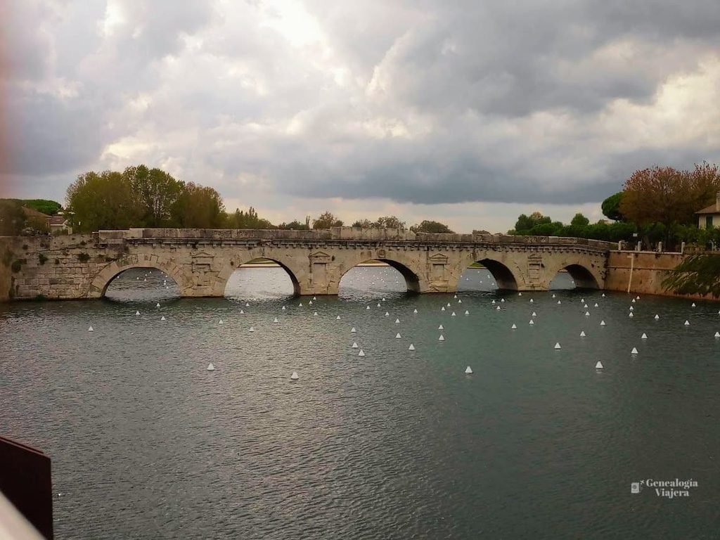 Vista panorámica del histórico Puente de Tiberio en Rímini, Italia, durante el mes de octubre. El puente de piedra con cinco arcos cruza un río donde flotan numerosas boyas blancas en forma de pirámide, bajo un cielo cubierto de densas nubes otoñales.
