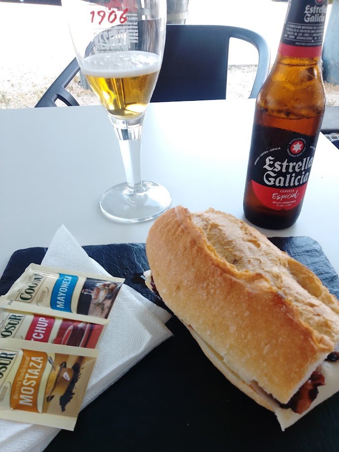 Un sándwich o bocadillo en pan rústico servido sobre una tabla de piedra negra junto a una botella y una copa de cerveza Estrella Galicia.
