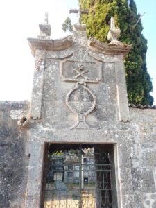 Antigua puerta de piedra granítica que da acceso al cementerio parroquial de Melón, Galicia, coronada por relieves barrocos de una calavera con huesos cruzados y cruces de piedra.
