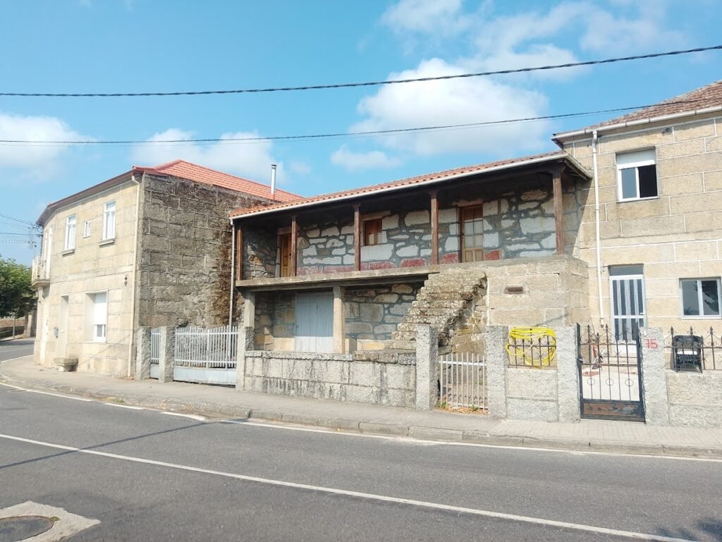 Casa tradicional gallega de piedra con escaleras exteriores y un largo balcón de madera en la planta alta, situada junto a la ruta bajo un cielo azul con nubes