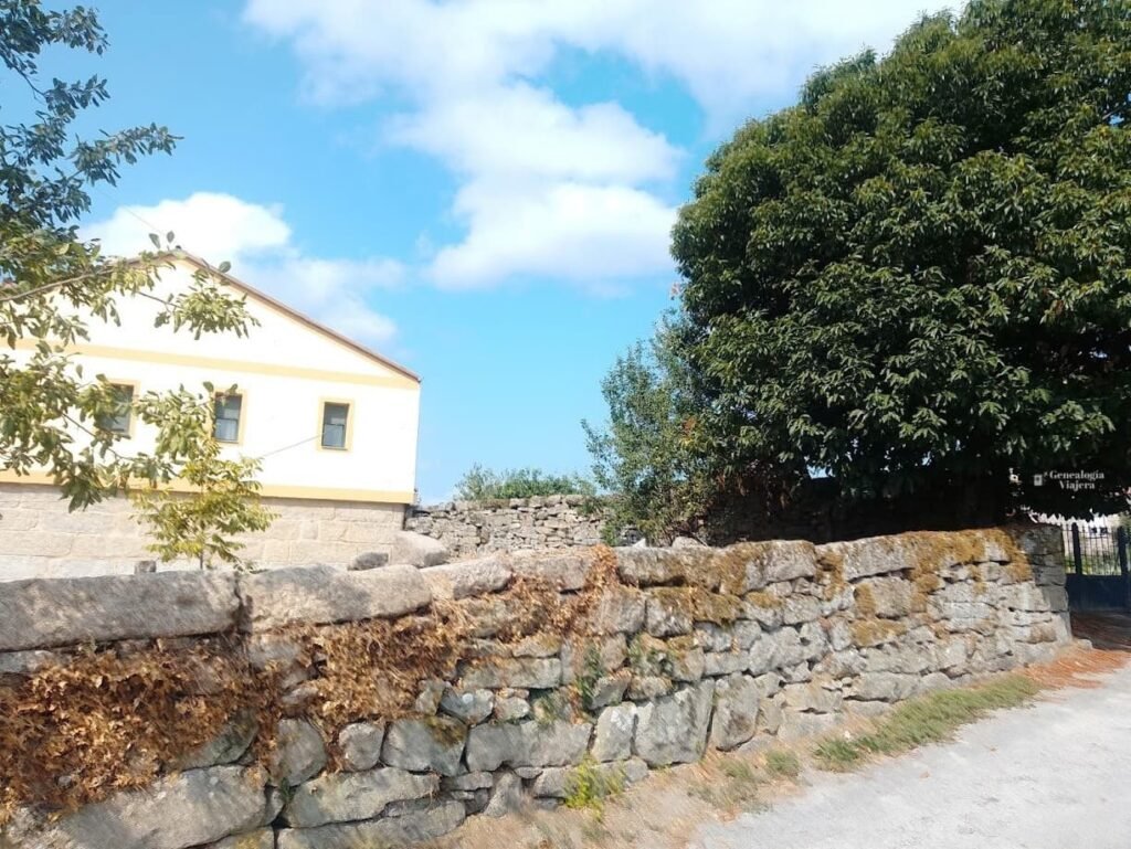 Vista de una calle asfaltada en Vilar de Santos flanqueada por un largo muro rústico de piedra gallega y casas tradicionales con tejados de tejas.