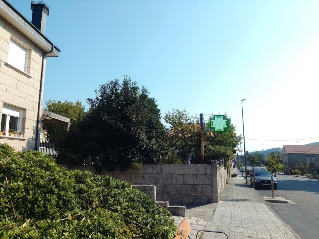 Calle pavimentada en el pueblo de Melón con un cartel luminoso verde de farmacia montado sobre una estructura metálica junto a un muro de piedra.