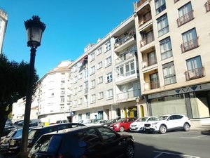 • Calle asfaltada en el centro urbano de O Carballiño con filas de autos estacionados y edificios de departamentos modernos con locales comerciales.
