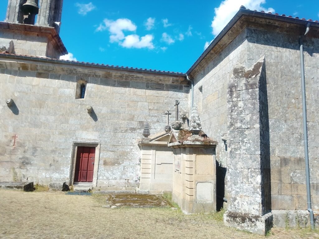 Vista de perfil de la Iglesia de Santa Baia de Banga en O Carballiño, destacando su torre campanario de piedra y los nichos del cementerio al fondo bajo un cielo despejado