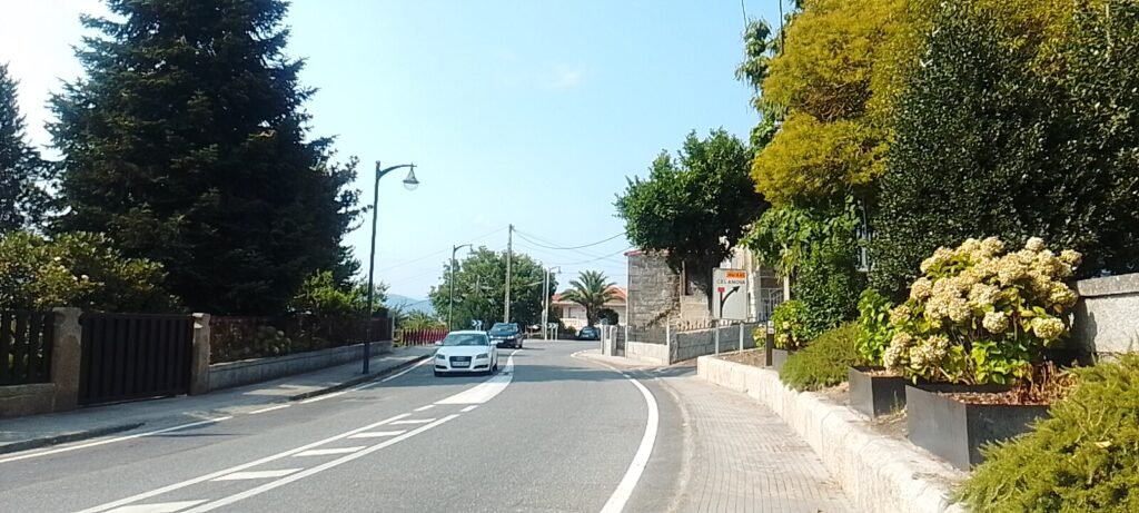 • Vista de la carretera principal en curva a su paso por Vilar de Santos, flanqueada por veredas con plantas, chalets residenciales, árboles grandes y un cartel indicativo hacia Celanova bajo un cielo despejado.