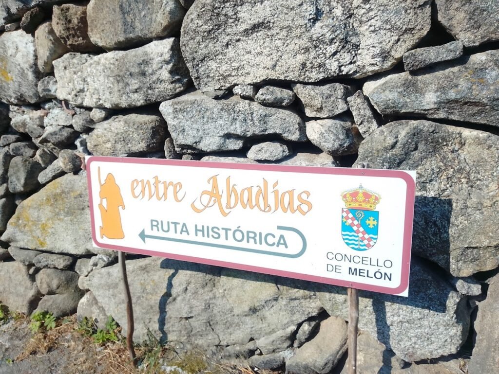 • Cartel alargado de color blanco con bordes granates que indica la dirección de la Ruta Histórica Entre Abadías en el Concello de Melón, Galicia, montado sobre un muro tradicional de grandes piedras rústicas.