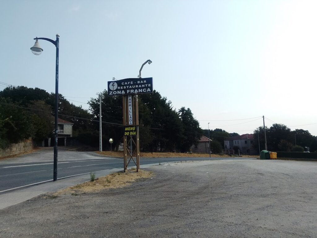 Cartel metálico elevado que anuncia el Café Bar Restaurante Zona Franca junto a una ruta bordeada de árboles en Vilar de Santos
