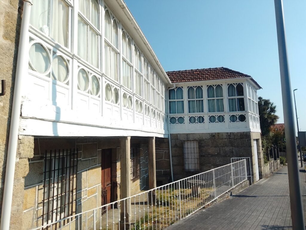 Fachada lateral de un edificio antiguo de piedra en Melón con grandes galerías tradicionales de madera blanca y cristales en el piso superior.