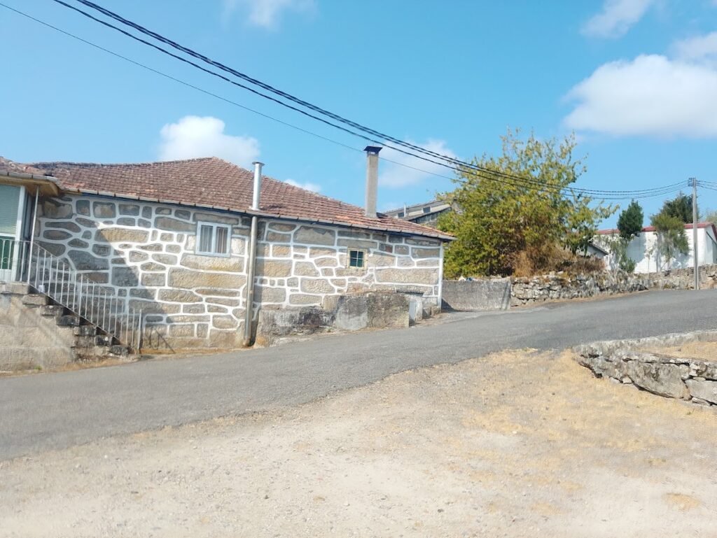 Casa de piedra típica gallega con escaleras exteriores y juntas de cemento blanco, ubicada junto a una calle en cuesta bajo un cielo azul con nubes.