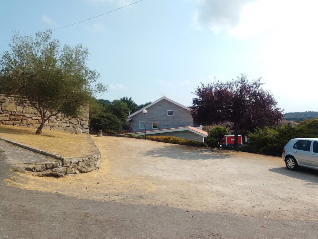 Una casa residencial moderna de piedra con tejado a dos aguas, rodeada de un gran terreno despejado, árboles y un auto gris estacionado a la derecha.