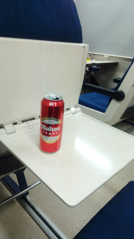 na lata de cerveza Mahou apoyada sobre la mesa plegable de plástico blanco de un asiento azul impecable dentro del tren interurbano español.