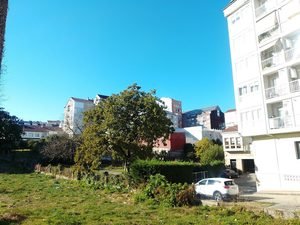 • Zona residencial en O Carballiño mostrando un terreno verde en primer plano y edificios modernos de viviendas bajo un cielo azul brillante.
