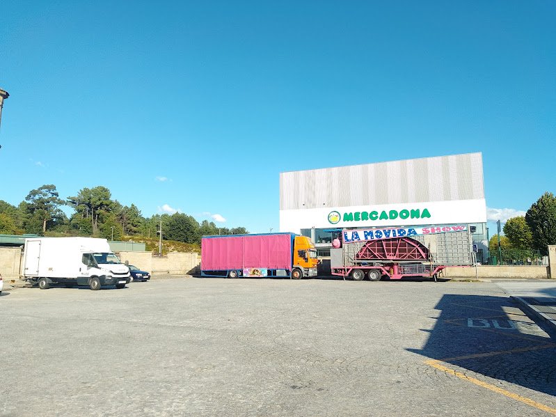 • Amplia playón asfaltado frente a la estación de O Carballiño con camiones de feria estacionados y el edificio del supermercado Mercadona de fondo bajo un cielo despejado.