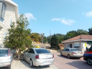Vista de la plaza junto al Ayuntamiento de Vilar de Santos, con autos estacionados en primer plano, un gran árbol a la izquierda y un edificio con paneles informativos al fondo bajo un cielo despejado.