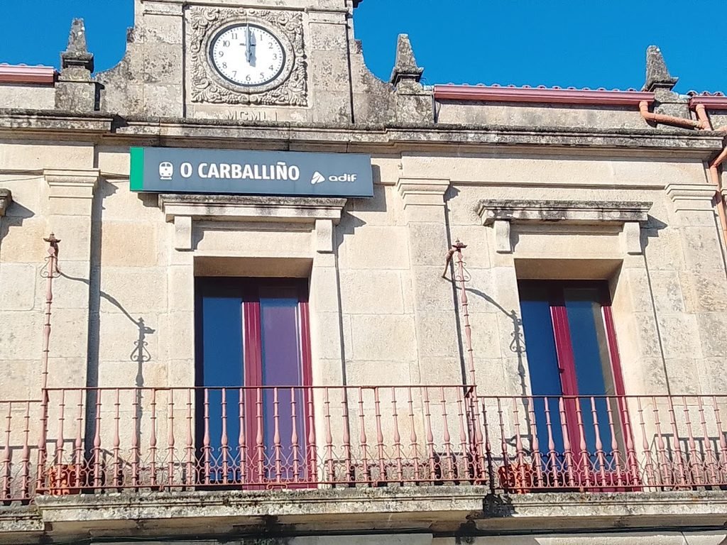 Detalle de la planta superior de piedra de la estación de tren de O Carballiño, con un balcón de hierro, el cartel verde oficial de Adif y un gran reloj antiguo en la parte superior.