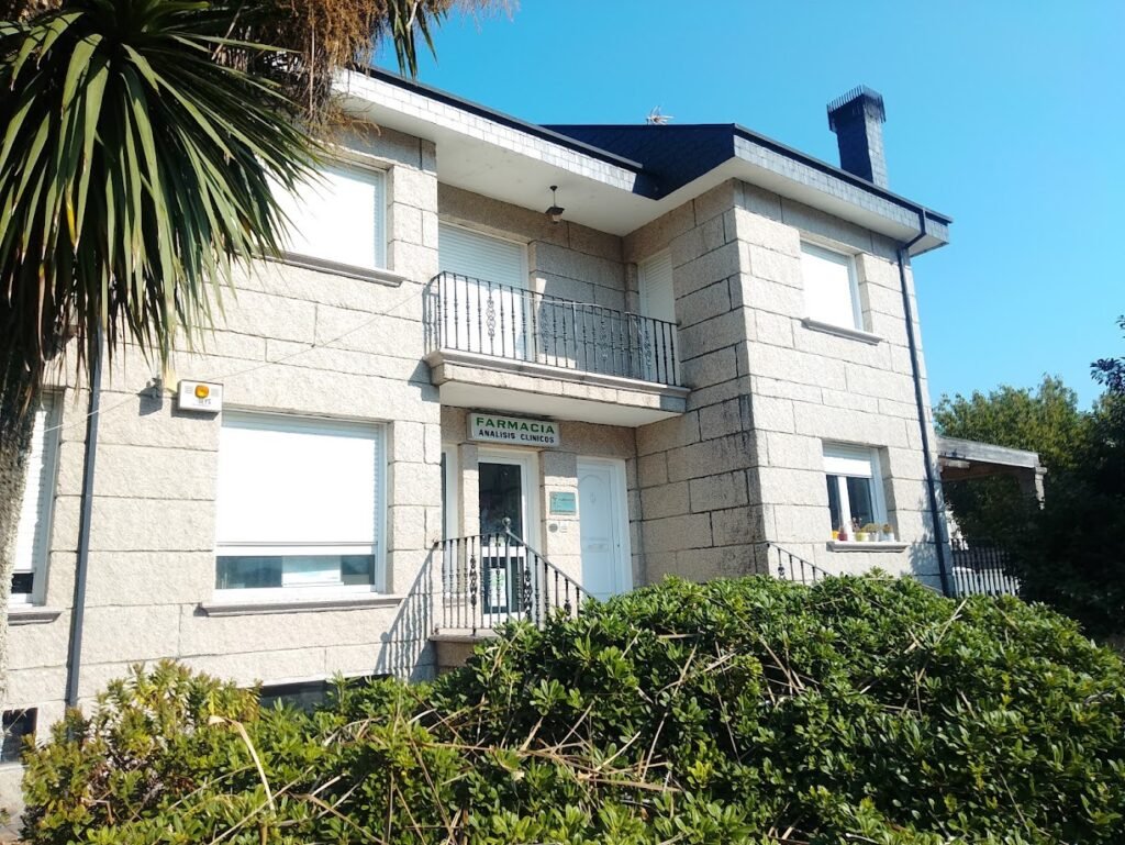 Edificio residencial y comercial construido en granito claro donde funciona la farmacia local de Melón, Galicia, con plantas verdes en el frente.