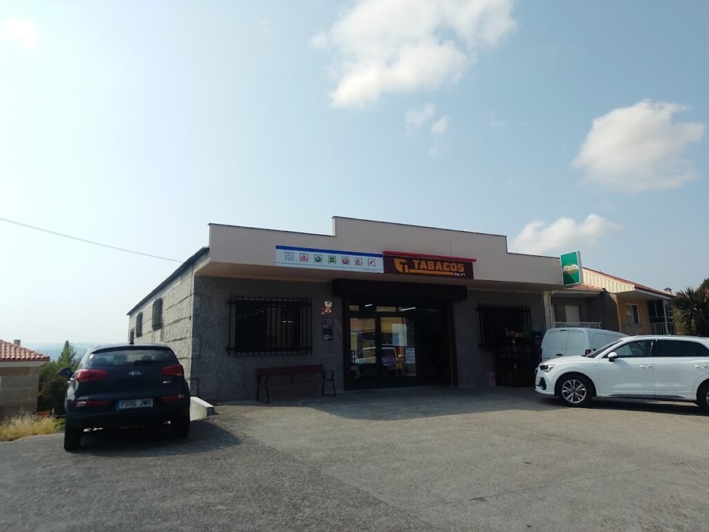 • Edificio comercial a pie de carretera en Vilar de Santos que alberga el Bar Restaurante Zona Franca y un estanco con el cartel oficial de Tabacos, con autos estacionados al frente bajo un cielo azul con nubes.
