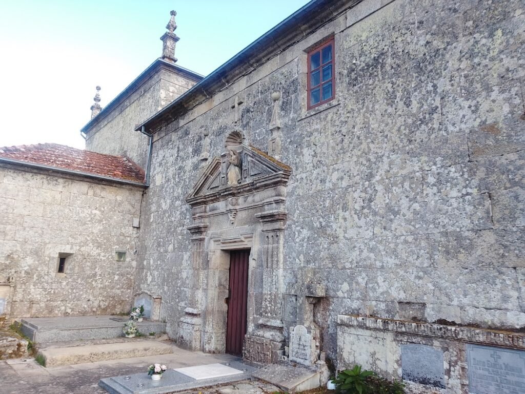 Muro exterior de sillería de piedra de la iglesia de Santiago de Corneda con una puerta de madera oscura, una pequeña ventana superior y sepulturas de granito en la base