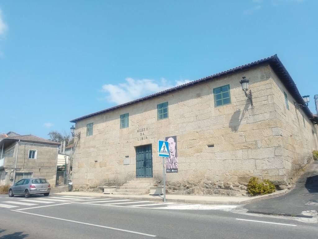 Vista de la fachada de piedra del Museo da Limia en Vilar de Santos, una edificación histórica de dos plantas con ventanas verdes, escalinatas de acceso y un cartel con la imagen de Antón Tovar junto a la carretera.