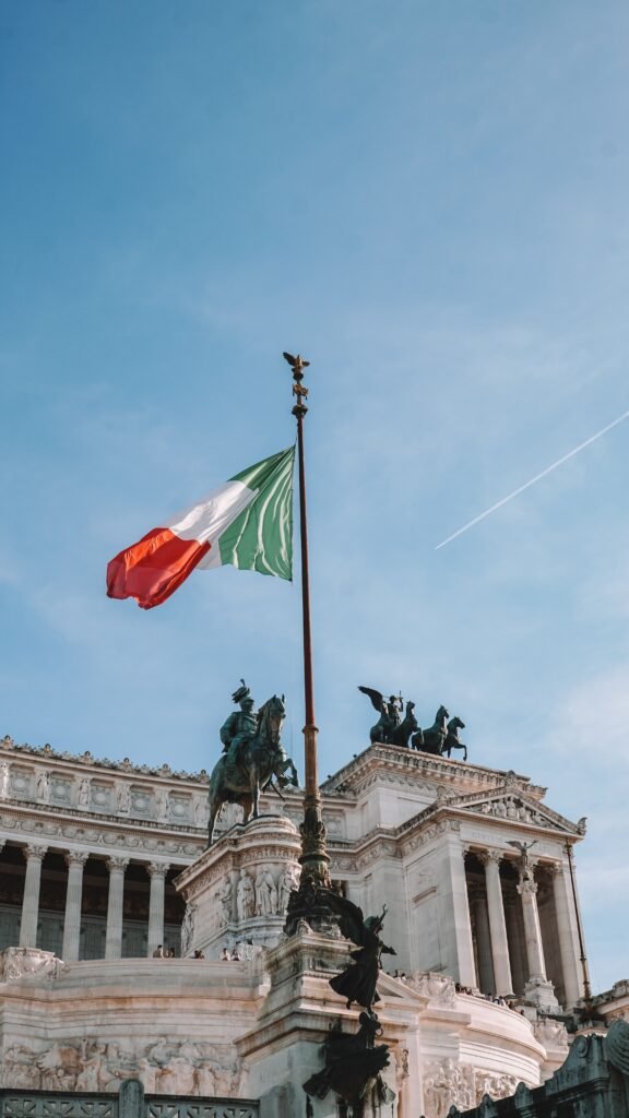 Monumento Vittoriano en Roma con la bandera de Italia flameando en primer plano bajo un cielo azul claro, seleccionado como portada para el artículo sobre la suspensión del referéndum de la ciudadanía italiana.