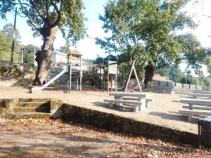 Parque infantil de madera y mesas de piedra para pícnic bajo árboles grandes en un entorno rural de Melón, Galicia.