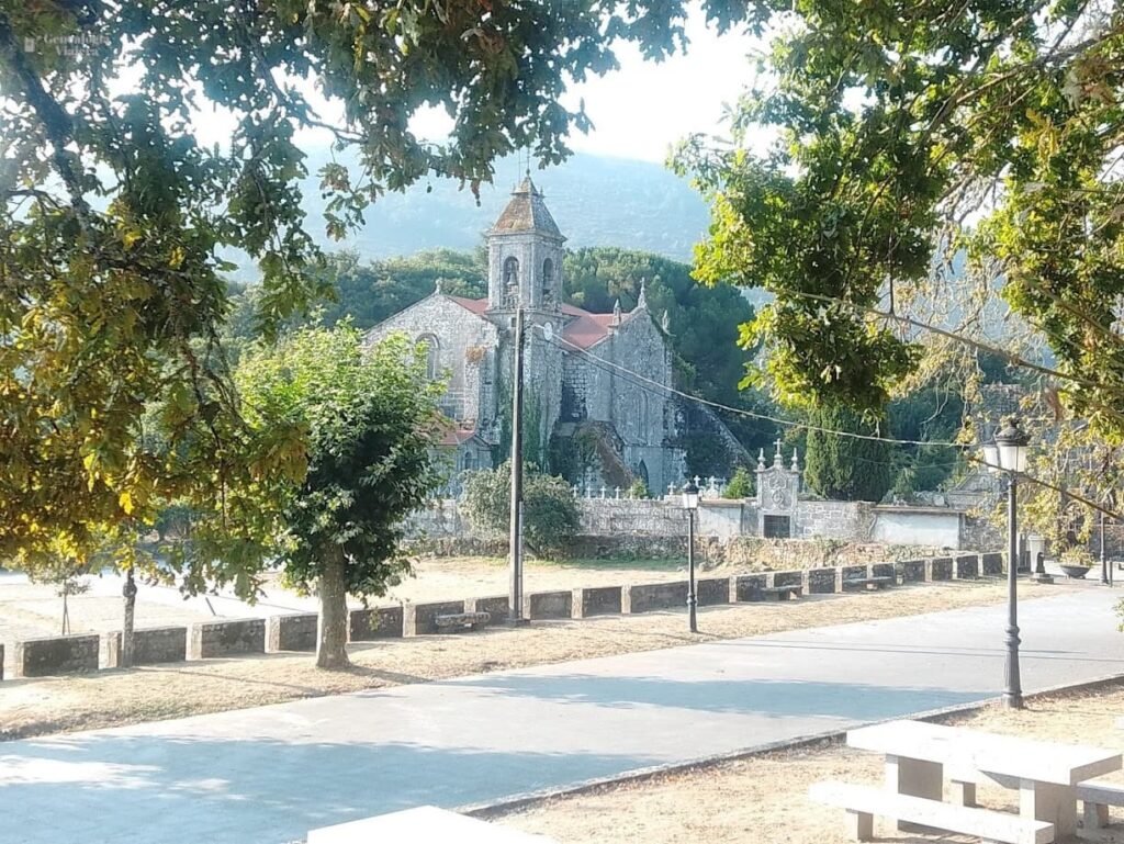 Camino pavimentado junto a una zona arbolada con bancos y mesas de piedra en Melón, Ourense, con la torre de la iglesia al fondo entre las hojas.
