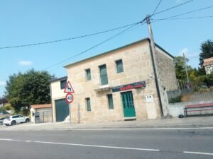 Fachada de una casa gallega de piedra en Vilar de Santos que alberga la Peluquería Noelia, situada junto a la ruta con una señal de tráfico de velocidad máxima de 40 kilómetros por hora en el frente.