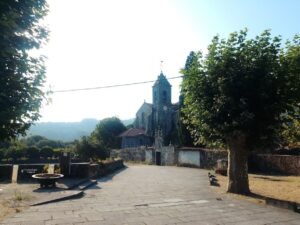 Amplia calle empedrada en Melón que conduce hacia el Monasterio de Santa María, rodeada de árboles verdes y muros rústicos bajo un cielo azul brillante.