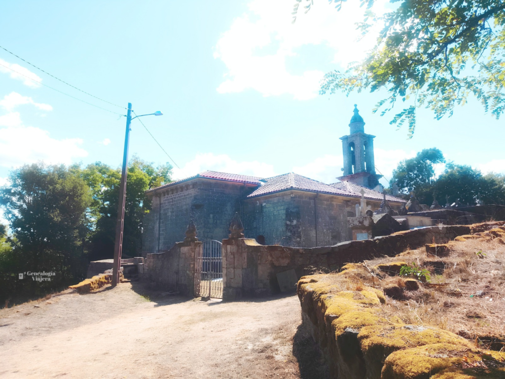 • Vista panorámica exterior de la Iglesia de Santa Baia de Banga bajo un cielo despejado, mostrando su estructura barroca de piedra, el campanario elevado y el muro perimetral con una reja de entrada sobre un camino de tierra.