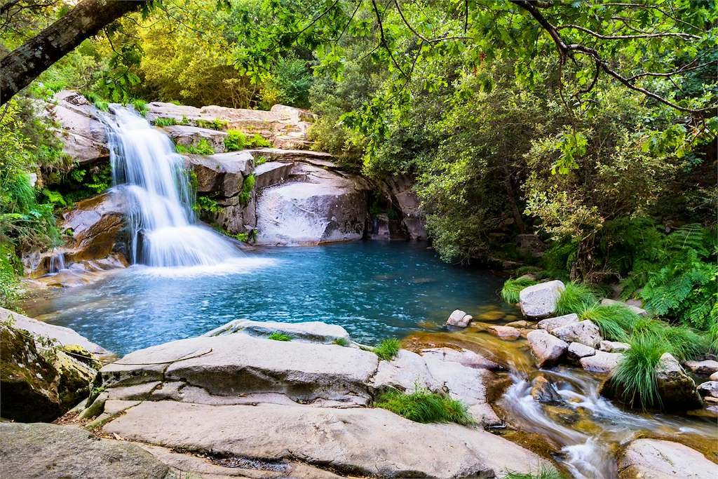 Cascada de agua cristalina cayendo sobre una gran piscina o poza natural de color turquesa rodeada de grandes rocas graníticas y densa vegetación forestal verde en Melón, Galicia.